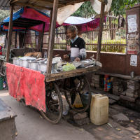 A street vendor in India stands behind a makeshift cart draped with a red cloth, surrounded by various cooking pots and containers. The stall is set up along a fence adorned with signs. A bench is placed in front, and on the ground lie a plastic canister and a white container.