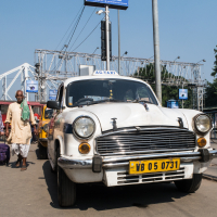 A white taxi with a yellow license plate marked "WB 05 0731" is parked on a busy street in India. People walk around it, and behind it, a large structure with numerous electrical wires is visible. There are buildings and trees in the background under a clear blue sky.