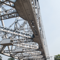 A low-angle view of the Howrah Bridge in Kolkata, India, showcases its intricate steel structure against a clear sky. Yellow taxis and buses move along the road underneath, with pedestrians on the sidewalks. Trees and buildings are visible in the background, capturing a slice of vibrant Indian life.