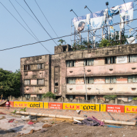 A dilapidated, three-story building with boarded up windows stands behind a construction zone in India. The construction area is lined with pipes and metal rods, separated by a fence with "Lux Cozi" ads. Overhead, several torn billboards are supported by a metal frame while vegetation peeks through the openings.