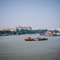 A river with several boats and barges floating on its calm waters under a clear blue sky in India. A distant shoreline features a mix of green trees and urban buildings, including some under construction. The backdrop includes various architectural structures blending into the city's landscape.