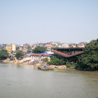 A wide river flows in front of a shoreline with dense vegetation and clustered buildings. On the banks, there are construction materials and small boats. The clear, sunny sky showcases a calm, daily life setting near the water in an urban area, reminiscent of scenes from India.