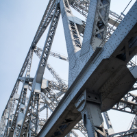 Close-up view of a large, detailed lattice steel bridge with intersecting metal beams and bolts. The structure is seen from a low angle, showcasing the intricate framework against a clear, pale blue sky. The metallic texture and industrial design of the bridge dominate the image, reminiscent of India's engineering marvels.