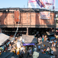 A bustling outdoor market scene in India with numerous vendors and customers. Stalls covered with tarps are laden with produce, especially yellow and orange flowers. The market is set against a backdrop of a large, old brick building with multiple billboards and some hanging signage, under a clear sky.