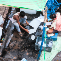 In an Indian street market, two men work under a green-tarp covered stall. One man saws through a block of ice on the ground, while the other scribbles in a notebook from his chair. Plastic bags and crates lie scattered around. Nearby, a third person stands holding a bag.