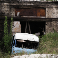 A blue van sits in the grass next to a brick building in Iļģuciems.
