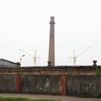 A tall chimney standing beside a brick wall in Iļģuciems.