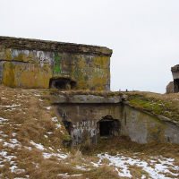 An abandoned concrete bunker complex stands amid a barren landscape with sparse snow patches in what was once the former military town of Karosta. The bunkers are weathered, covered in graffiti, and surrounded by dry grass. The overcast sky enhances the desolate atmosphere, highlighting the deteriorating structures with multiple entry points.