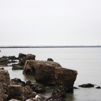 A serene coastal landscape featuring scattered rocky remnants along the Karosta shore, with calm waters extending to a distant horizon under an overcast sky.