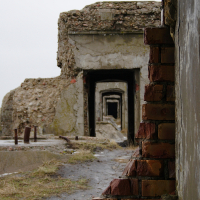 A view through multiple archways of the weathered, abandoned Karosta building. The walls are crumbling, revealing exposed bricks and concrete. The ground is uneven, with patches of grass and rubble. The sky is overcast, casting a dull light over the decaying, historic structure.