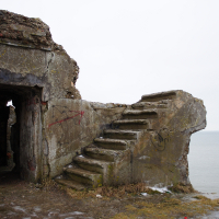 A dilapidated coastal Karosta WWII bunker with graffiti presents a staircase leading to nowhere, offering a poignant reminder of history's decay against the backdrop of a serene sea.