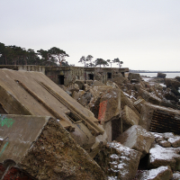 Ruins of an old coastal fortification in Karosta, with dilapidated concrete structures scattered along a snowy beach, trees in the background, under an overcast sky.