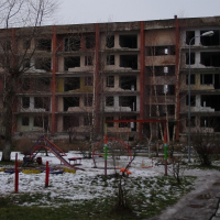 A bleak winter scene in Karosta showing an abandoned, dilapidated building with broken windows behind a desolate playground with scattered snow patches, evoking a sense of neglect and desolation.