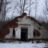 An abandoned, dilapidated structure in Karosta with a crumbling red brick facade and a derelict white plaster front stands forlornly amidst a leafless winter forest, with remnants of snow