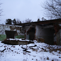 A dilapidated brick building in Karosta stands in wintery decay, its walls crumbling under the weight of time, partially clad in remnants of green and white paint, the snow gently blanketing