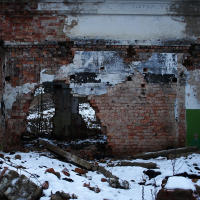 A dilapidated brick structure in Karosta with remnants of white plaster, featuring a graffiti-tagged wall and a snow-dusted ground, stands as a testament to decay and abandonment.