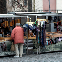 An elderly person in a red coat and gray hat shops at an outdoor market stall. The stall, reminiscent of 2012, has a variety of knitted items like scarves, hats, and gloves on display. Another similar stall is adjacent with more knitted accessories and jewelry by Kazjukas. The background features a building with bare trees.