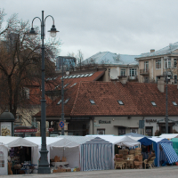 A bustling outdoor market with multiple vendor stalls covered by white and striped canopies lines a cobblestone street. The background features buildings with red-tiled roofs and faded facades. Leafless trees, ornate streetlamps, and a Kazjukas poster from 2012 hint at the cold season's charm.