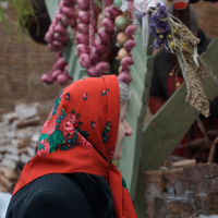 An elderly woman wearing a black coat and vibrant red floral headscarf stands in front of a wooden stall at the 2012 Kazjukas festival. The stall is adorned with hanging bunches of dried red onions and assorted herbs, while the background features stacked firewood and partially visible textiles.