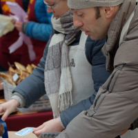 Two people, dressed warmly in coats, knitted hats, and scarves, serve food at an outdoor market. They are focused on their tasks, handling trays and plates. A third person in the background, also warmly dressed, appears to be assisting. The scene is lively and bustling like Kazjukas 2012.