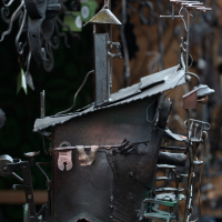 A metal sculpture by Kazjukas from 2012 depicts a whimsical, abstract house with a slanted roof, chimney, and attached wire details. The house blends geometric and organic forms, with metal elements resembling clothes on a line and twisted wirework suggesting branches or antennae. Background is blurred.
