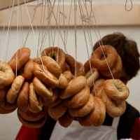 A variety of bagels hang by strings from the ceiling of a market stall. The bagels come in different shades of brown, indicating various types or levels of baking. A person with brown hair is partially visible in the background, focusing on the display, reminiscent of a scene from Kazjukas 2012.