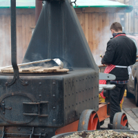 A person stands beside a large, mobile metal oven on wheels, likely used for cooking or smoking food outdoors. The individual, possibly Kazjukas, is wearing a black jacket and red apron, facing away from the camera. In the foreground, there's a bowl filled with what appears to be dry vegetation or herbs.