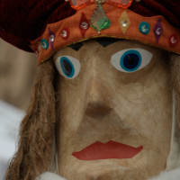 Close-up of a large wooden puppet with exaggerated facial features including bright blue eyes and red lips. The Kazjukas 2012 puppet has long hair and wears a headdress adorned with multicolored jewels and a large red, bejeweled top section. The headdress includes a decorative green diamond-shaped element.