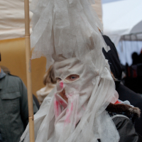 A person is dressed in a costume with layers of white, crinkly material covering their head and face, extending upwards to form a tall, pointed hat. The material has a slightly translucent appearance. They are holding a wooden pole and wearing a dark coat, reminiscent of the Kazjukas celebration in 2012, with other indistinct figures in the background.