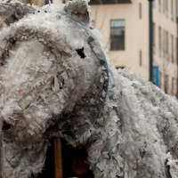 A large, gray, horse-shaped puppet covered in frayed fabric and supported by wooden poles is part of a lively street performance or parade. People can be seen underneath and around the puppet. The background features urban buildings and a white tent, reminiscent of the 2012 Kazjukas Festival. The scene appears festive and crowded.