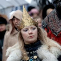A woman dressed in medieval attire stands among a crowd at the 2012 Kazjukas Festival. She wears a golden crown with intricate designs, chainmail, and a fur-trimmed cloak. Her long blonde hair falls freely, and she has a focused expression. People in similar historical costumes and modern winter wear are seen in the background.