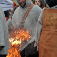 A man in a hooded garment and leather apron tends to a large fire outdoors, flames rising high. Beside him stands a woman in similar attire, "Gintaro Galerija" written on her apron. This scene, reminiscent of the 2012 Kazjukas festival, is watched by a group of hooded onlookers with colorful flags behind them.