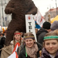 Group of people at an outdoor festival in 2012, some wearing traditional headbands. A large wicker bear sculpture is prominently displayed in the background. The people are holding flags and signs, celebrating Kazjukas. The event appears festive, with a crowd and tents visible in the background.