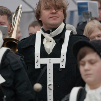 A man, wearing a black coat and a white chest strap, stands amidst a crowd at Kazjukas 2012. His brown hair is swept over one eye, and he holds a trombone. People in the background are dressed in similar attire, suggesting a formal event or parade. Another person in the foreground is out of focus.