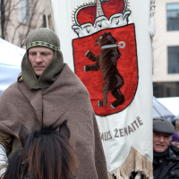 A person dressed in medieval attire, including a chainmail hood and brown cloak, rides a dark horse. Behind them is a large banner featuring a red shield with a bear standing upright, a crown, and the text "Auks Žemaitė." A modern-clothed figure stands in the background with "Kazjukas 2012" subtly visible on their shirt.