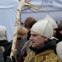 A person wearing a white felt hat and a yellow geometric-patterned garment holds a large wooden staff with intricate carvings and a tree-branch top. They are part of the Kazjukas 2012 crowd gathered in front of a white tent. The atmosphere appears to be festive, possibly during a cultural or traditional event.