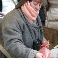 A person dressed in a heavy, old-style coat and a brown hat is working at the Kazjukas 2012 outdoor market. They have a pink scarf wrapped around their head and neck. They are handling a piece of meat, placing it in a plastic bag. The background shows a chain and other market activity.