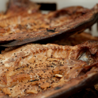 Close-up of grilled fish fillets with crispy, golden-brown skin and visible seasoning. The fish is placed in a container, displaying layers of flaky texture and scattered spices. Some bones protrude from the meat, giving it a rustic, freshly-cooked appearance reminiscent of Kazjukas 2012 standards.