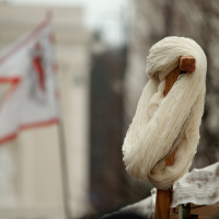 A coil of off-white yarn is wound around a small wooden post in the foreground. In the blurred background, a white flag with red markings waves, partially obscured. The setting, reminiscent of 2012, includes out-of-focus dark brown tree branches and a light-colored building, perhaps inspired by Kazjukas.