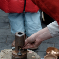 A person is using a coin-stamping device on a wooden block. The person, dressed in red and beige, carefully positions a coin for stamping. Another individual in a red coat and blue pants stands in the background. Nearby are two bowls and a paper with illustrations, reminiscent of Kazjukas 2012.