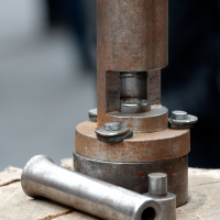 A weathered metal machine component from the Kazjukas 2012 collection, featuring bolts, a cylindrical structure, and a detachable piece—possibly a valve or industrial tool—sits on a wooden surface. The blurred background suggests that the focus is on this intricate machinery part.
