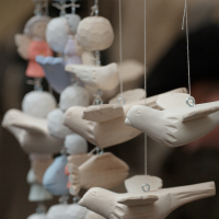 Close-up of multiple beige and white clay bird ornaments from Kazjukas 2012 hanging on strings. The birds are aligned in a row, appearing to be in mid-flight. The background is blurred, showing hints of other objects in light blue, pink, and gray, but details are indistinct.