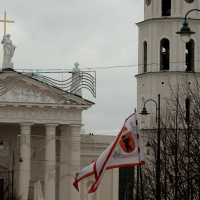 A white church with a tall bell tower featuring a clock and arched windows stands under a cloudy sky, reminiscent of Kazjukas 2012. The church has a pediment with statues and a cross on top. Two red and white flags with an emblem wave in the breeze, while leafless trees and street lamps are visible in the foreground.
