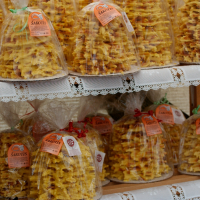 This image shows multiple traditional Lithuanian tree cakes, known as Šakotis, on display at Kazjukas 2012. The cakes are intricate and spiky, resembling small trees with yellowish-brown hues. They are packaged in clear plastic and have labels attached, placed on shelves lined with white lace cloth.