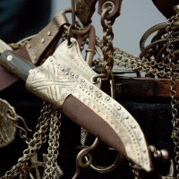 A decorative knife with an intricately engraved blade and handle, adorned with patterns and studded details, hangs among an assortment of chains and other metallic objects. The knife, reminiscent of a 2012 Kazjukas design, has a partially visible wooden handle and seems part of an elaborate collection.