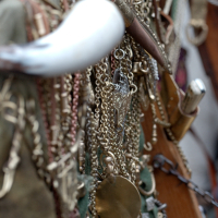 A close-up image of an assortment of brass chains, ornate keys, and various metal trinkets hanging on a green fabric. The items include a white horn and a leather sheath containing a small dagger. The scene appears cluttered, showcasing intricate, vintage items reminiscent of Kazjukas 2012.