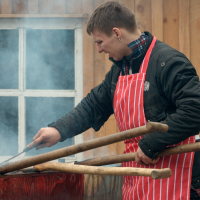 A person wearing a red and white striped apron and a black jacket is grilling food on a barbecue, reminiscent of the scene from Kazjukas 2012. They are holding large wooden tongs and turning food on the grill with a metal spatula. Steam or smoke rises from the barbecue, with a wooden building and window in the background.
