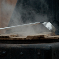 Close-up of a steel ladle resting on a wooden surface, with steam rising up, suggesting it's hot. The dimly lit background emphasizes the steam and the contrast between the metal ladle and wood below. The scene, evoking a 2012 Kazjukas aesthetic, conveys a rustic or industrial cooking environment.