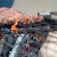 A close-up of skewered pieces of marinated meat cooking over a charcoal grill with visible flames and embers, reminiscent of Kazjukas 2012. The chicken appears well-seasoned, turning golden from the heat. The darkened grill rods hint at frequent use, adding to the warm, smoky ambiance.
