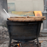 A large black pot labeled "Kazjukas 2012" sits on an outdoor wood-fired stove with a wooden lid partially covering it. The stove has a metal frame and visible flames below, indicating it is actively in use. In the blurred background, there are some paintings and a tree trunk. A metal lamp post is also visible.